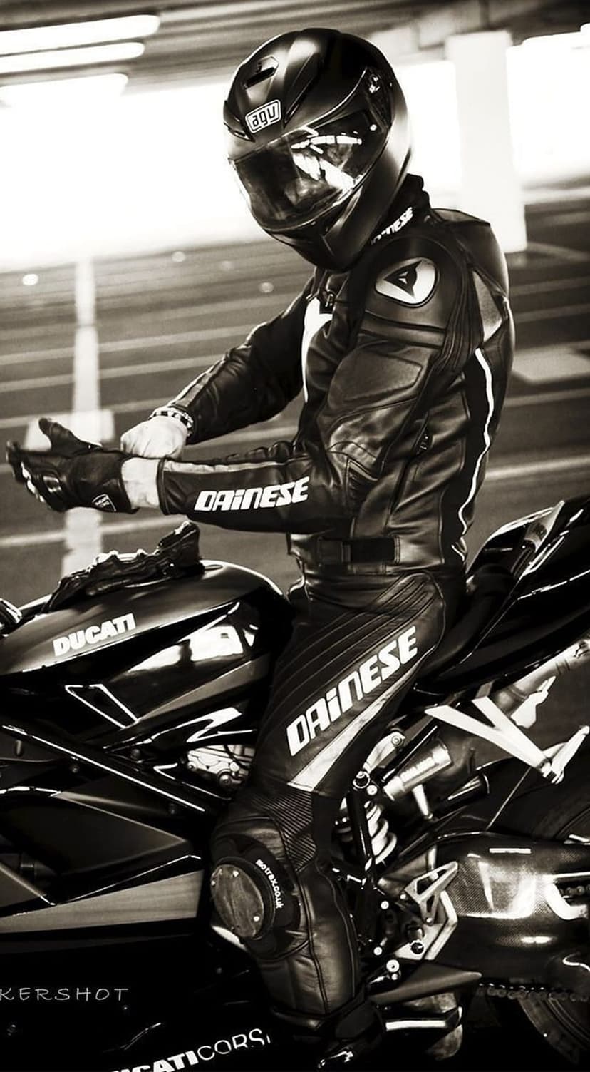 Biker in Dainese racing gear seated on a Ducati motorcycle, adjusting leather gloves in a high-contrast B&W view.