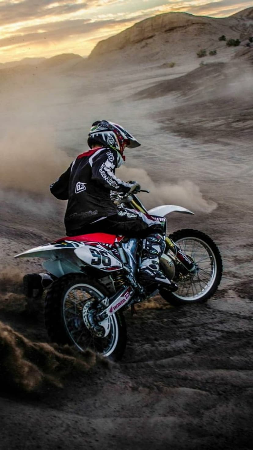 Motocross rider on a dirt bike creates a massive dust cloud in a sandy desert during sunset with long shadows.