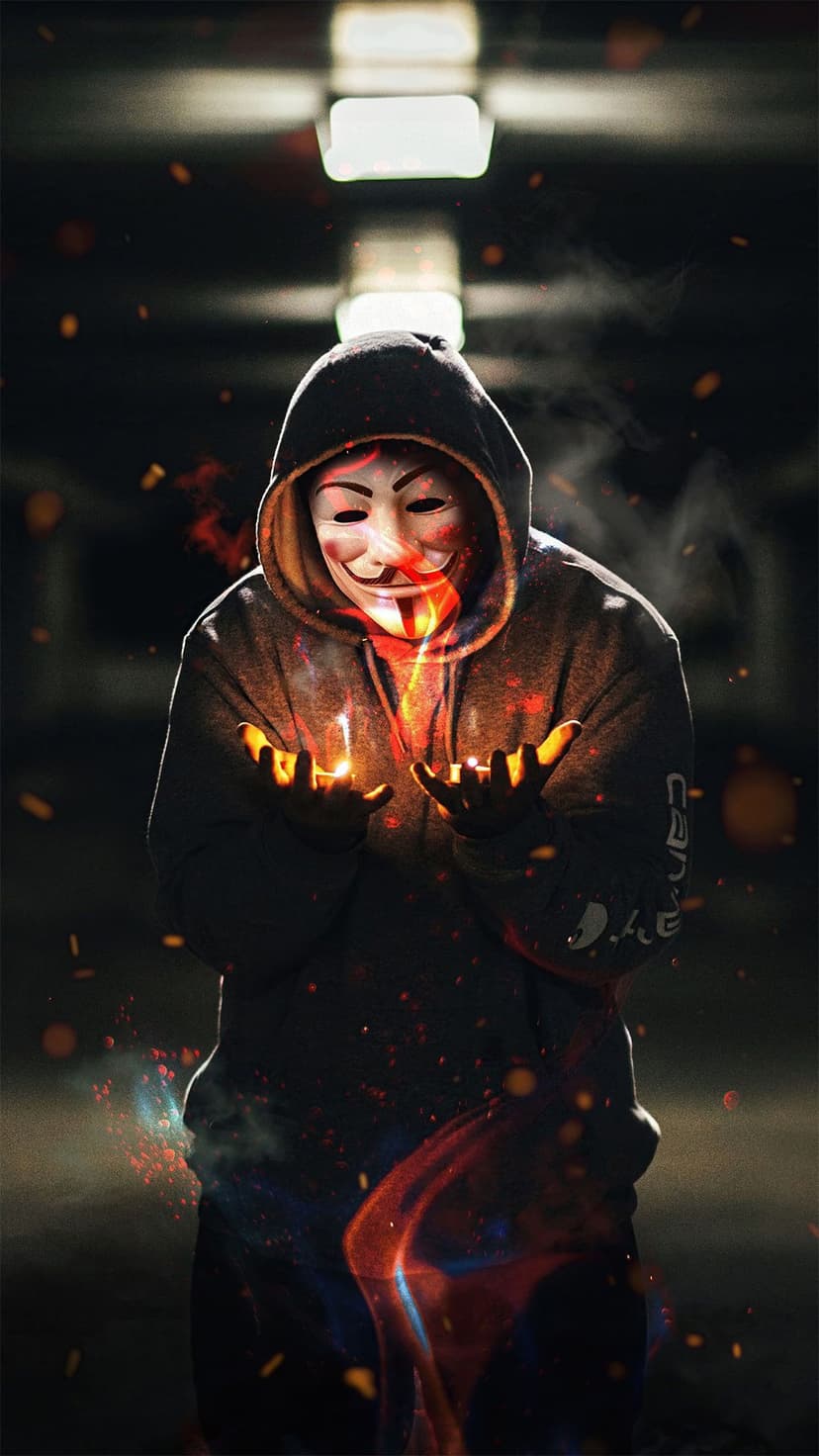 Masked person in a black hoodie holding a glowing ball of fire and orange sparks against a dark background.