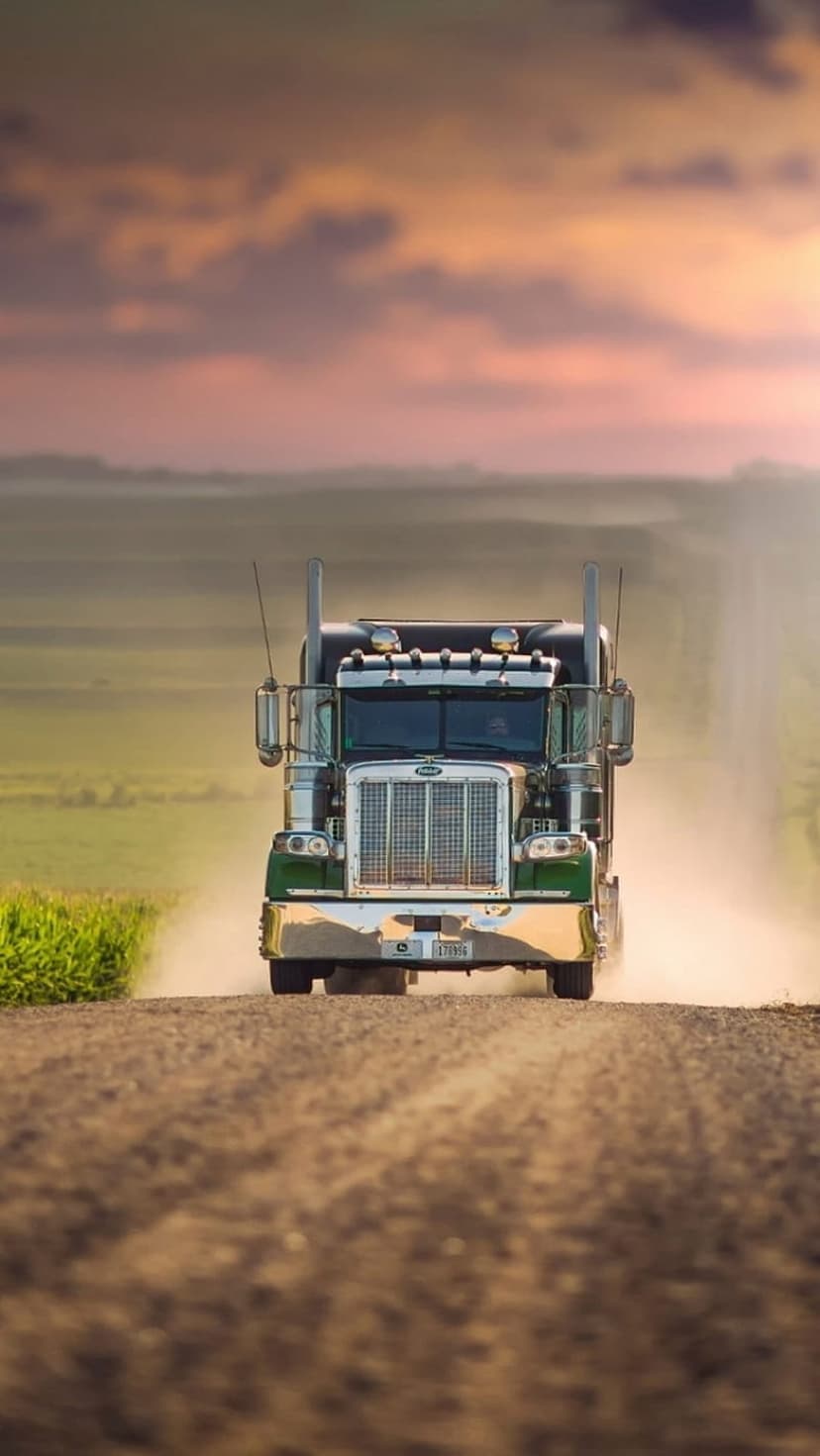 Green semi-truck driving on a dirt road at sunset with orange clouds, chrome details, and trailing dust clouds.