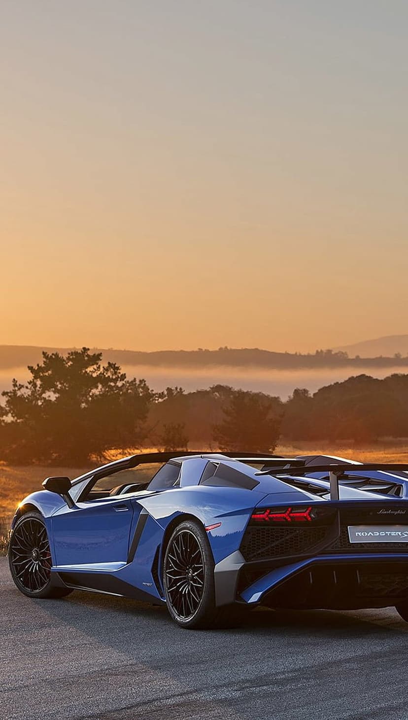Blue Lamborghini Aventador SV Roadster on a scenic road with a sunrise sky, rolling hills, and lush green trees.