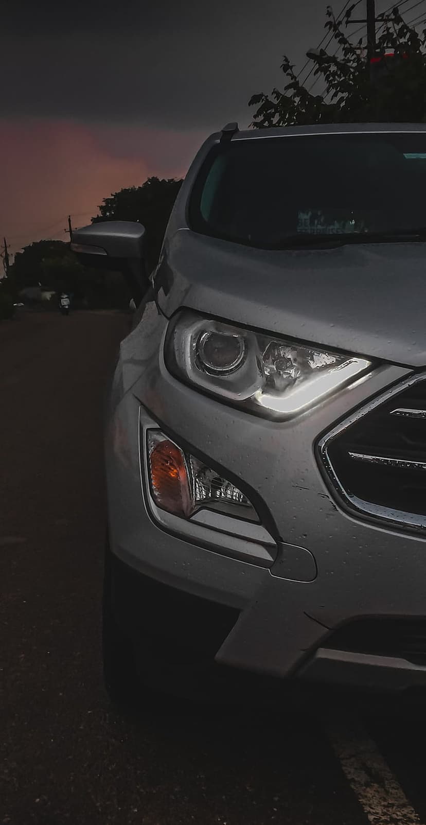 Ford EcoSport LED headlight and silver grille glowing against a moody pink and orange twilight sky at dusk.