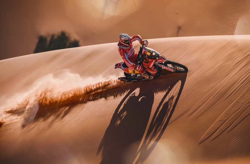 Motorcyclist in red gear leans into a sandy turn on a desert dune, kicking up a large dust cloud in harsh light.