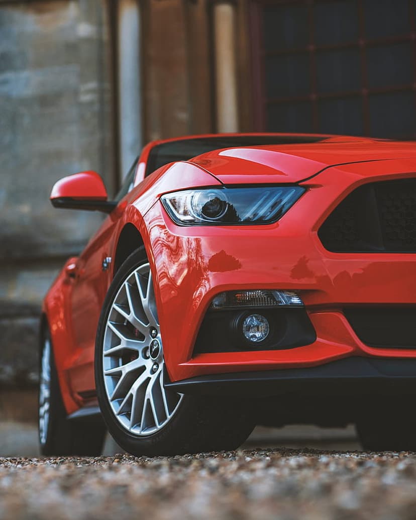 Red Ford Mustang front view with aggressive headlights and alloy wheels on a textured gravel and concrete street.