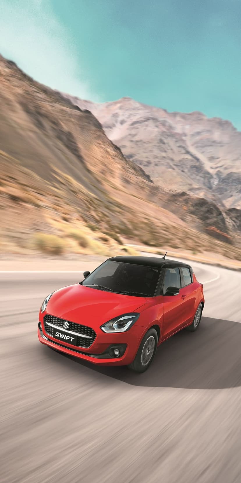 Red Suzuki Swift with black roof driving on a winding mountain road with motion blur and sunlit rocky peaks.