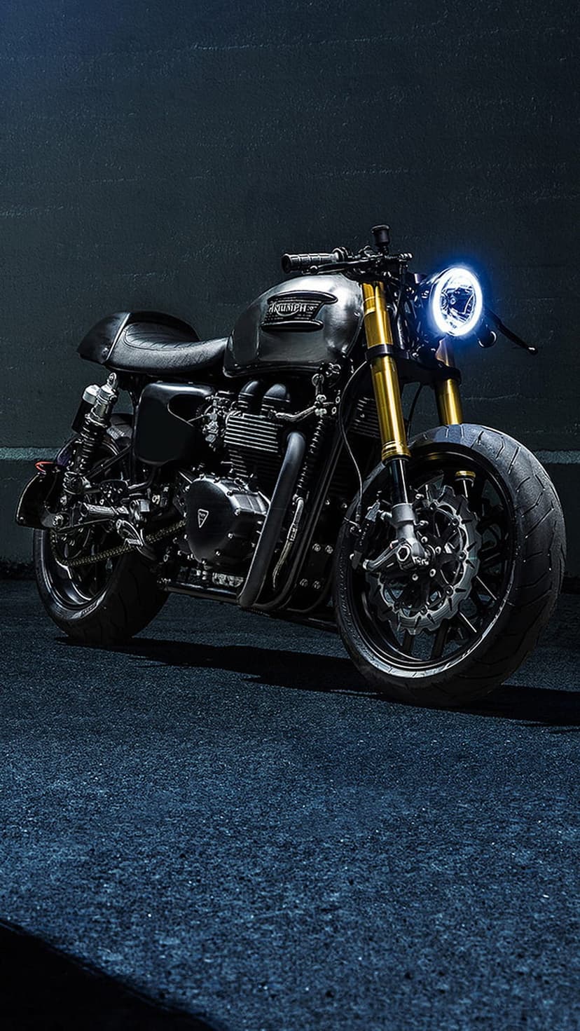 Silver Triumph cafe racer motorcycle with a bright LED headlight and golden forks on a dark textured background.