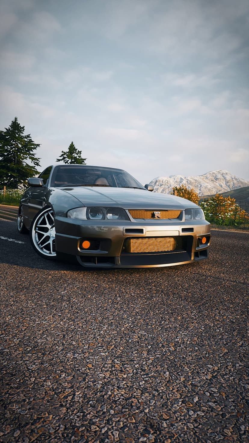 Nissan Skyline GT-R R33 in grey drives on a mountain asphalt road beneath a cloudy sky at a low camera angle.