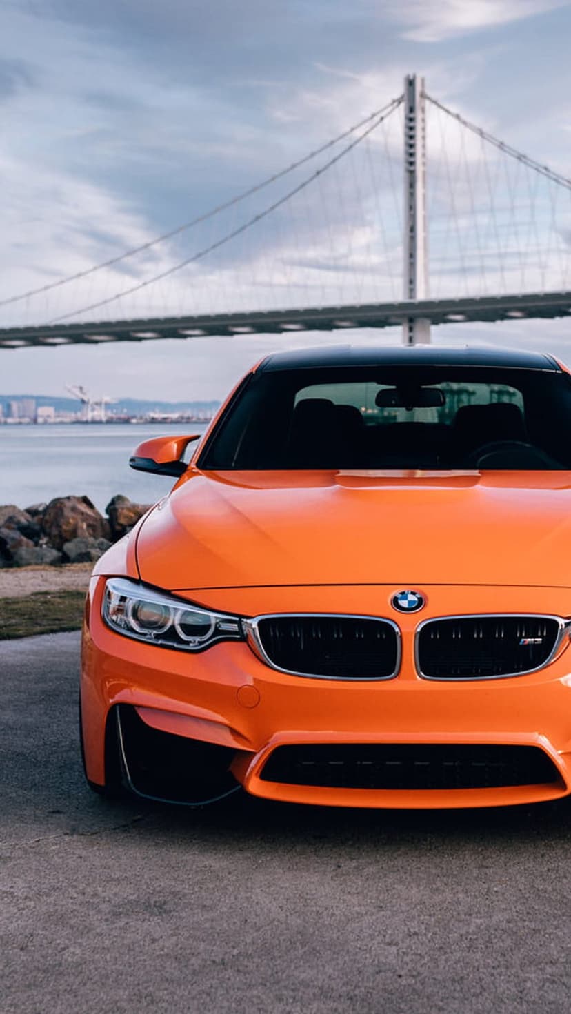 Orange BMW M4 parked on a coastal road with a large suspension bridge and blue ocean in the background.