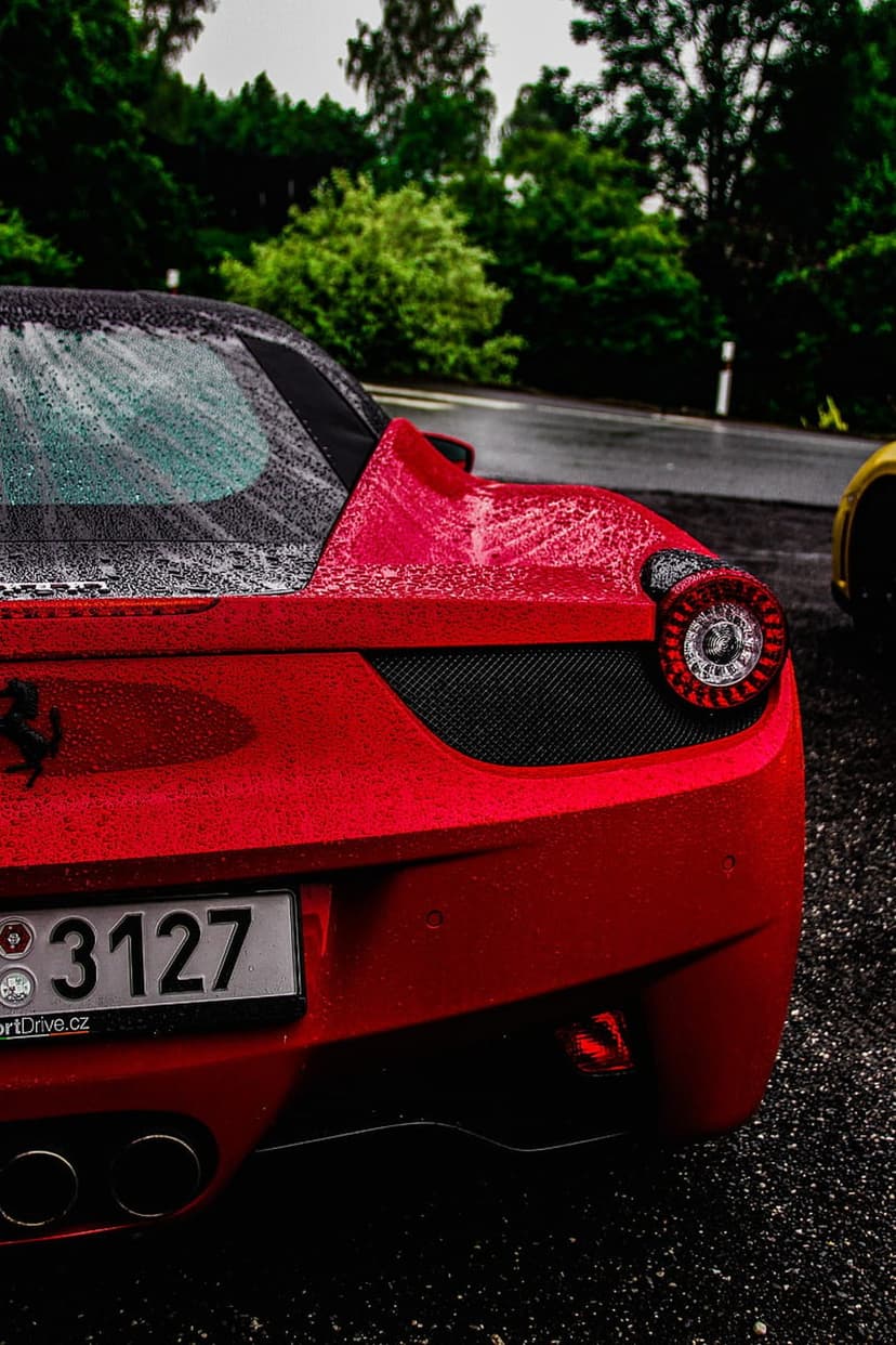 Ferrari 458 Italia rear showing red paint, water droplets, round taillight, and exhaust pipes on a wet road.