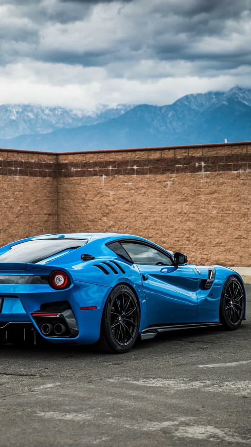 Blue Ferrari F12 TDF parked by a brick wall with cloudy mountain peaks in the background during daytime.