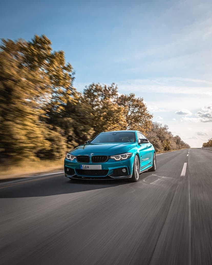 Blue BMW M4 coupe speeds along a scenic highway flanked by blurred orange autumn trees under a bright sky.