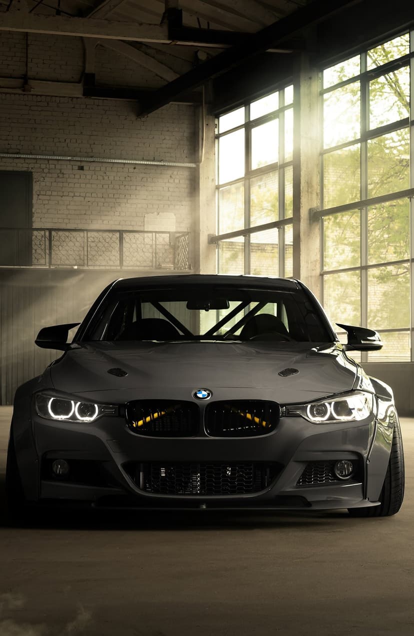 Matte black BMW M3 with a roll cage and widebody kit parked in a sun-drenched industrial warehouse.