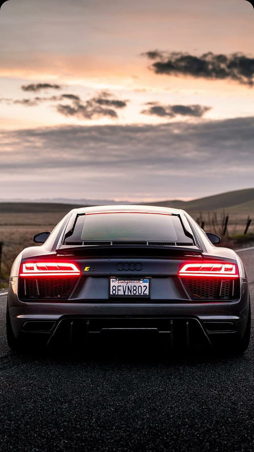 Audi R8 rear view with glowing red taillights parked on a dark road under a dramatic orange and purple sunset sky.