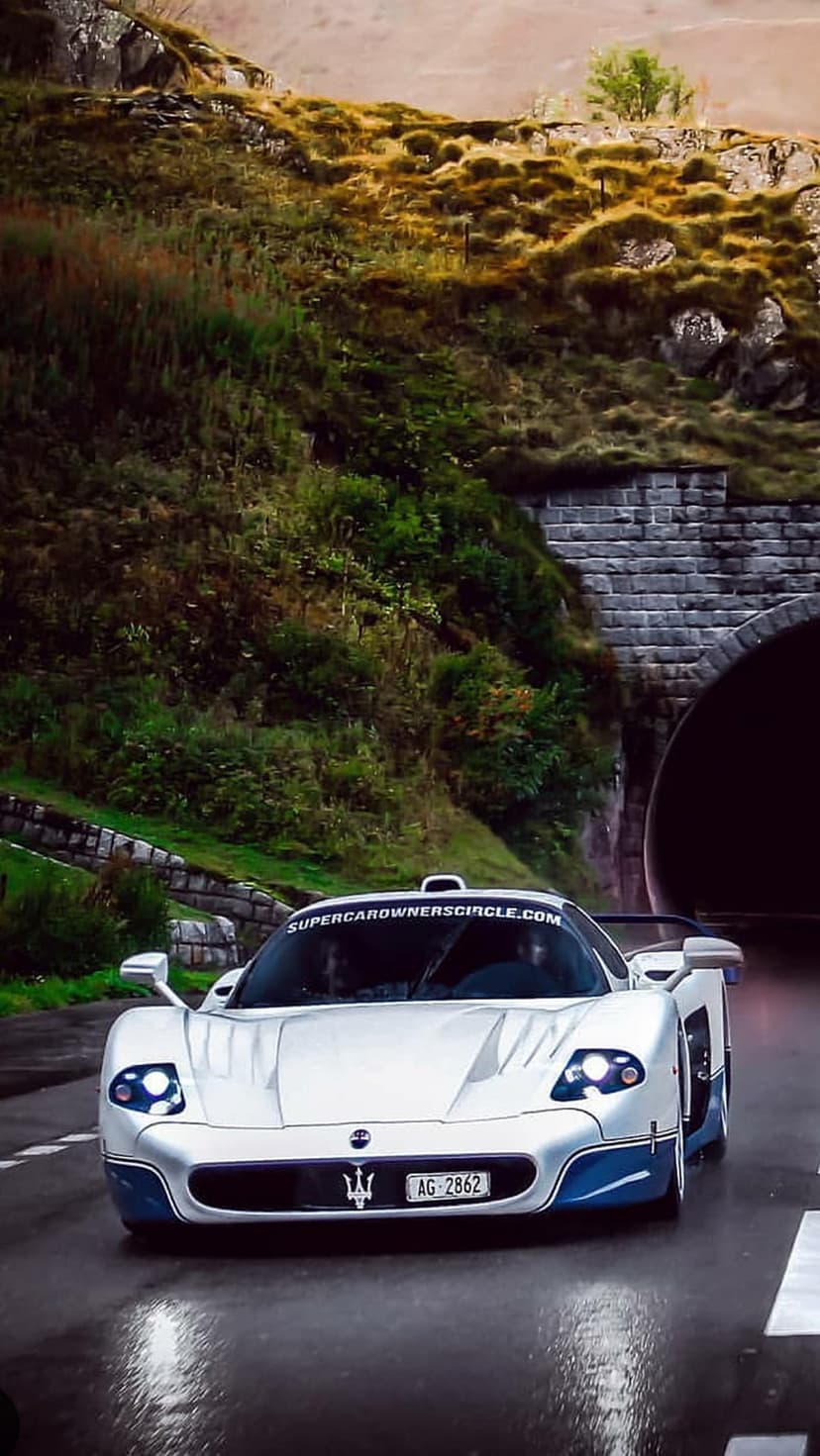 White Maserati MC12 supercar exiting a dark stone tunnel onto a wet road surrounded by green mountain slopes.