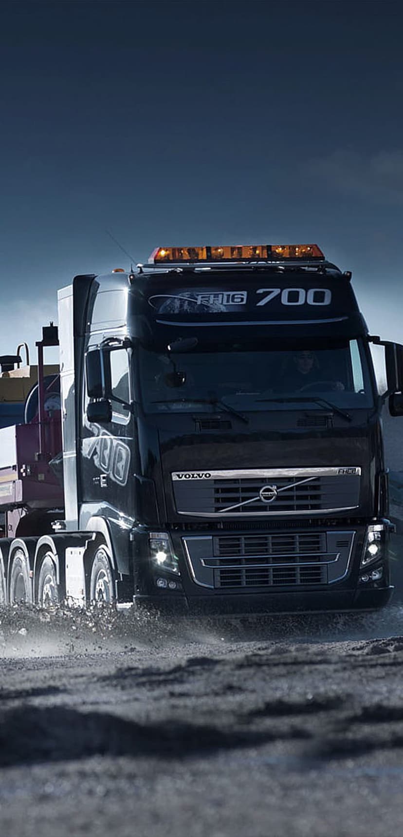 Volvo FH16 700 truck forging through a muddy puddle with water splashing around its massive wheels and amber lights.