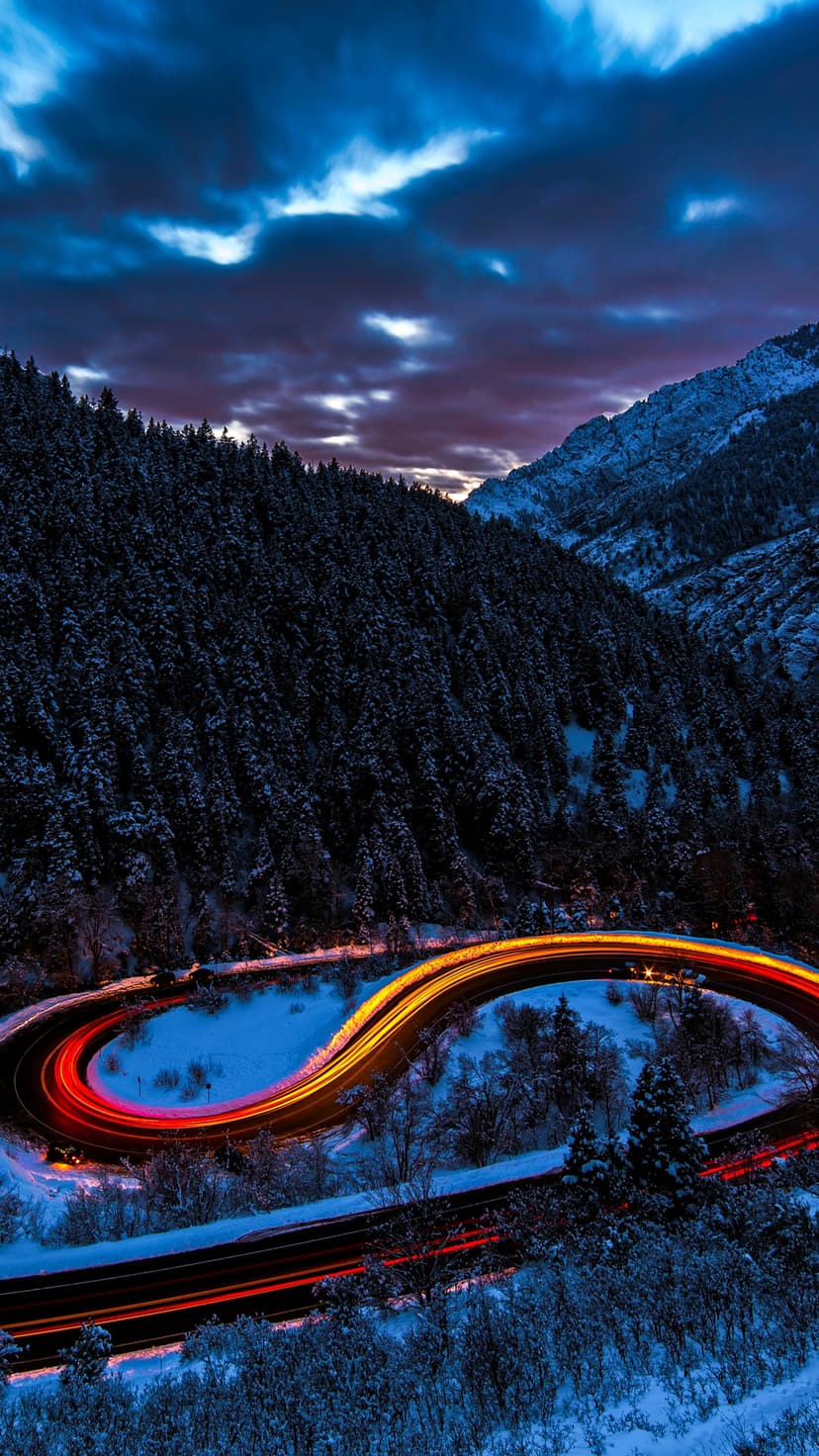 Orange and red light streaks curve along a snow-covered mountain road surrounded by dark evergreen trees.