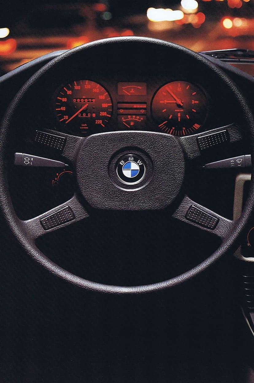 Vintage BMW steering wheel and glowing red dashboard gauges illuminated against a dark night city backdrop.