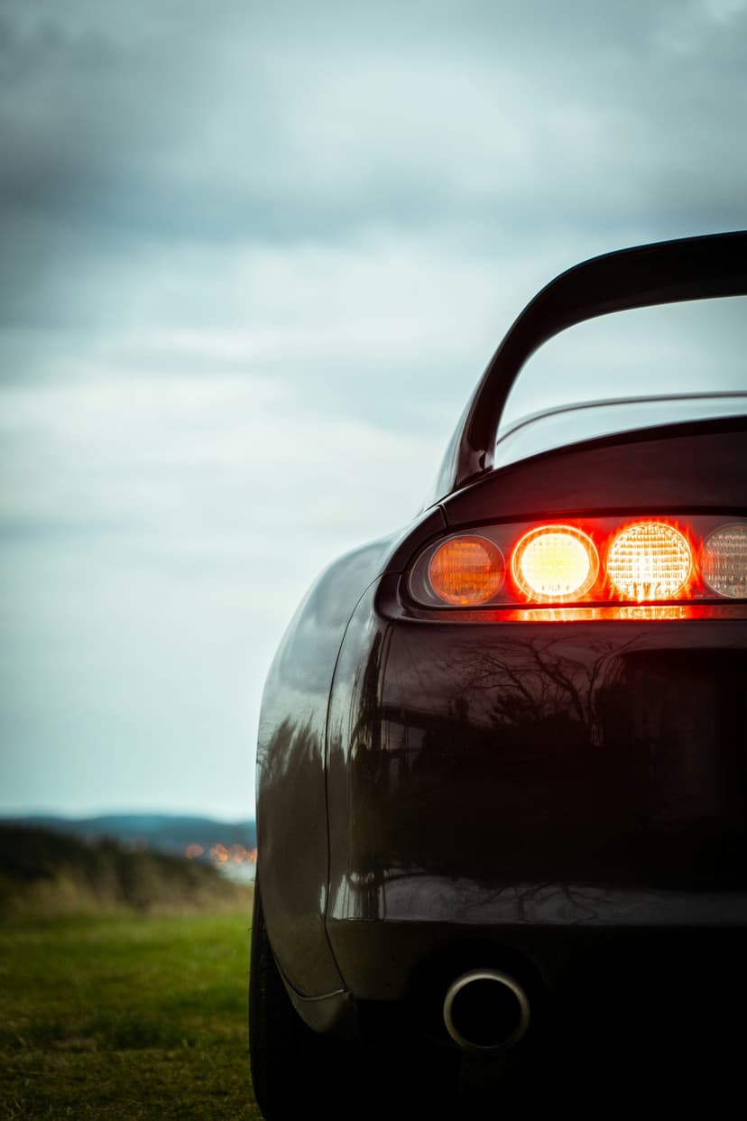Red sports car rear with illuminated circular taillights parked on grass against a cloudy twilight sky.