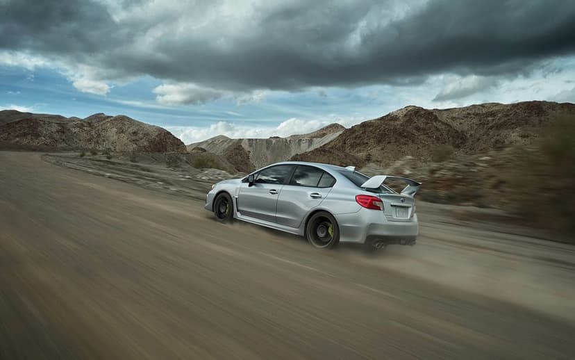 Silver Subaru WRX STI driving fast on a dusty desert road under dark dramatic clouds with mountain peaks.