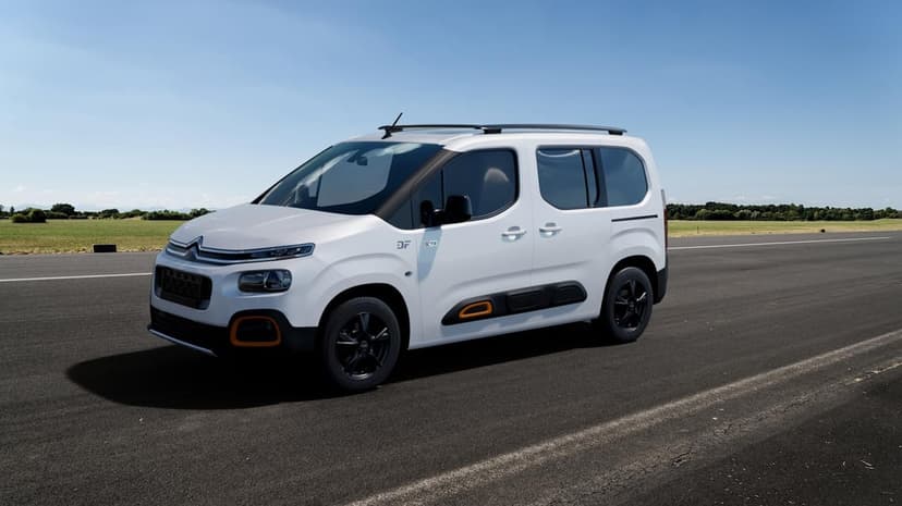 White Citroën van with orange accents on a grey asphalt road under a clear blue sky with distant green fields.