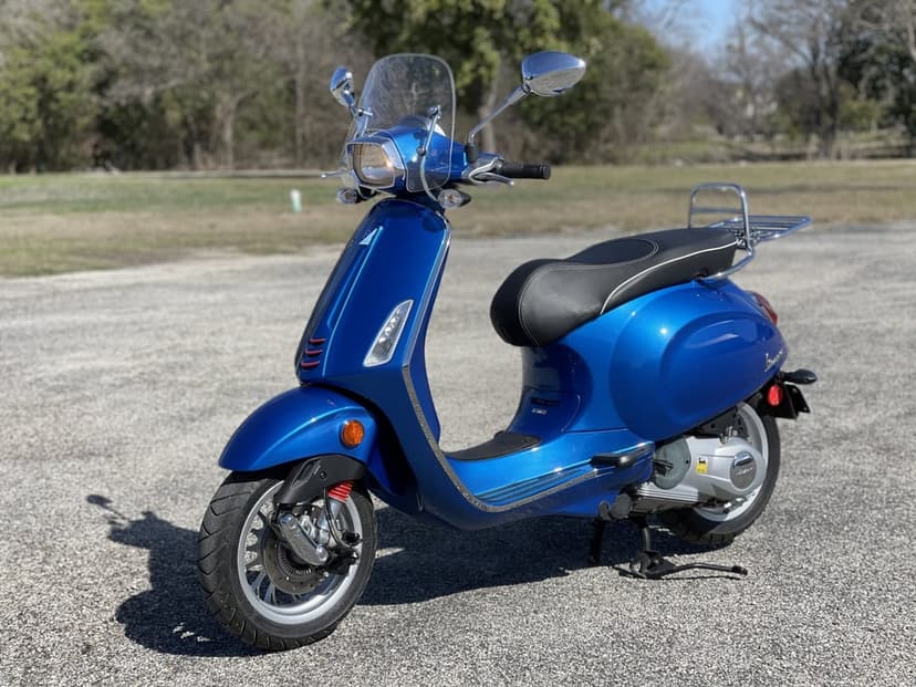 Metallic blue Vespa scooter with chrome accents parked on asphalt under bright daylight with a blurred background.