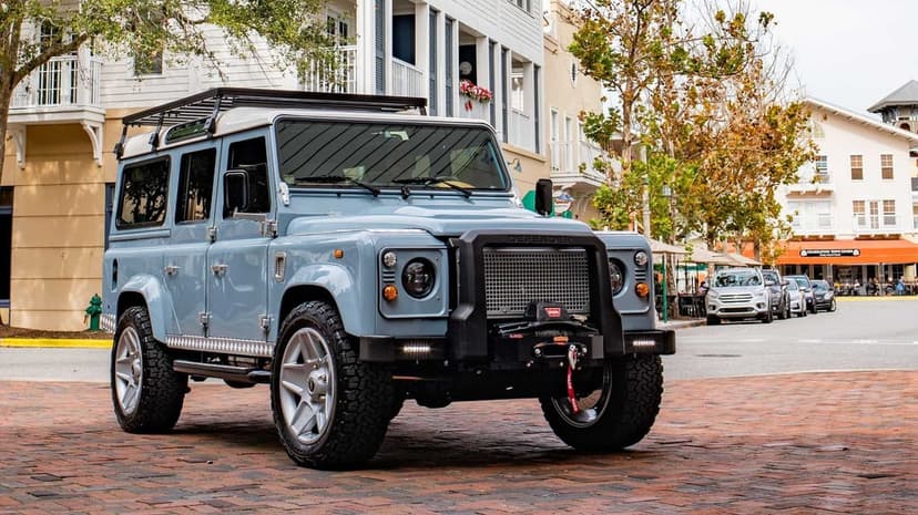Light blue Land Rover Defender with a black bull bar and roof rack on a cobblestone urban street.