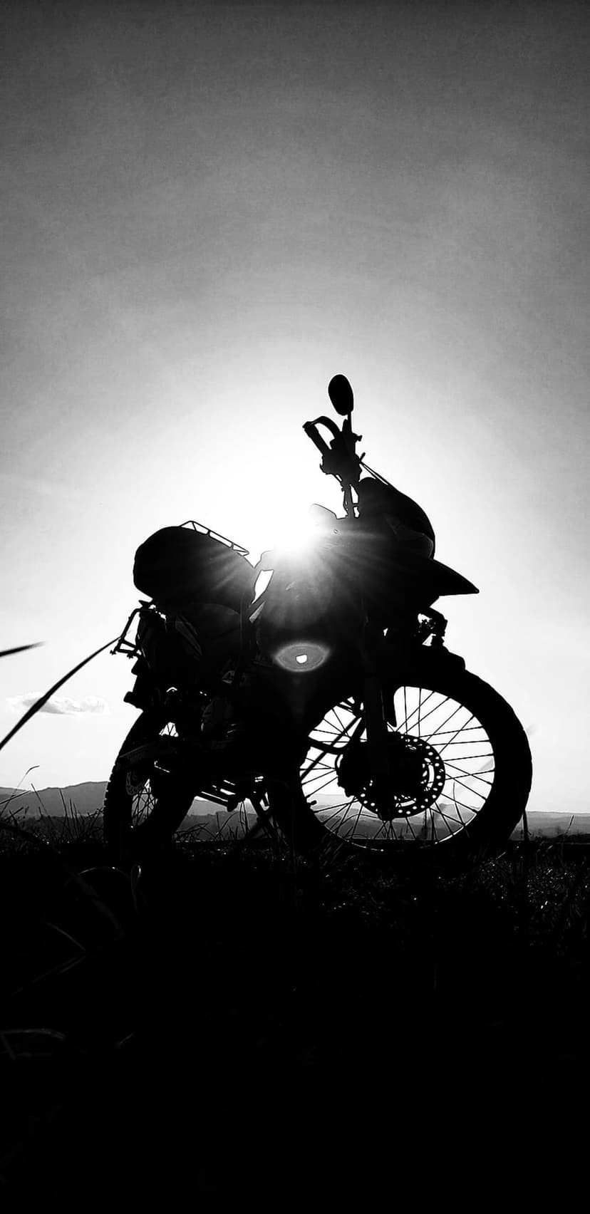 Motorcycle silhouette on a grassy hill with a bright sun flare and distant mountains in black and white.