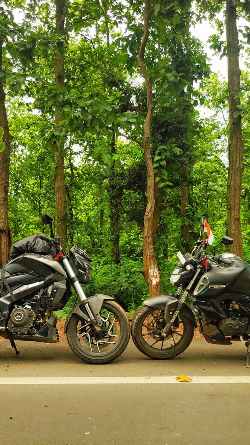 Two sport motorcycles park on a tree-lined asphalt road with an Indian flag attached to the handlebars.