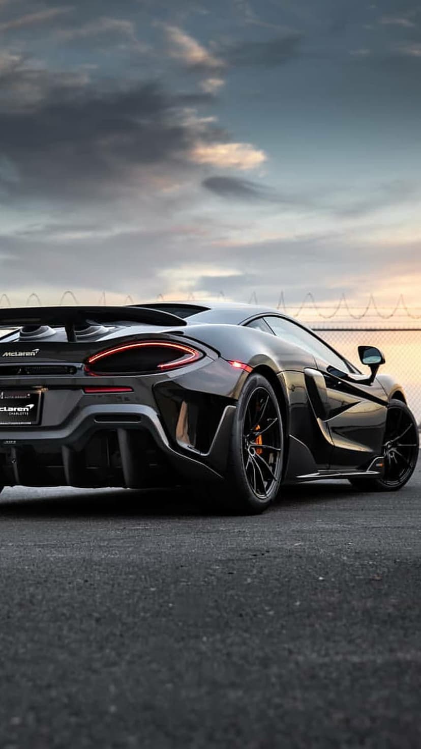 Black McLaren 570S supercar rear view with glowing red LED taillights on asphalt against a twilight sky.