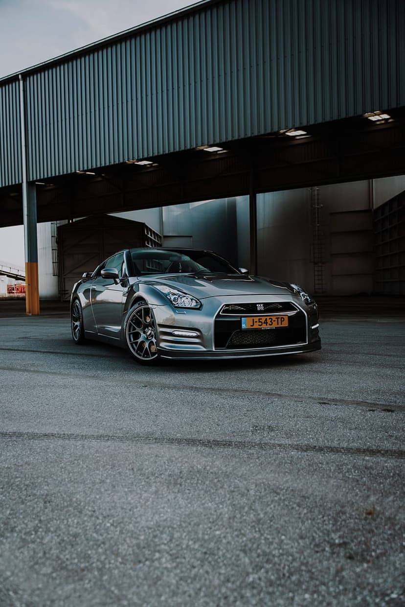 Silver Nissan GT-R R35 parked on asphalt in front of a corrugated metal industrial building during daylight.