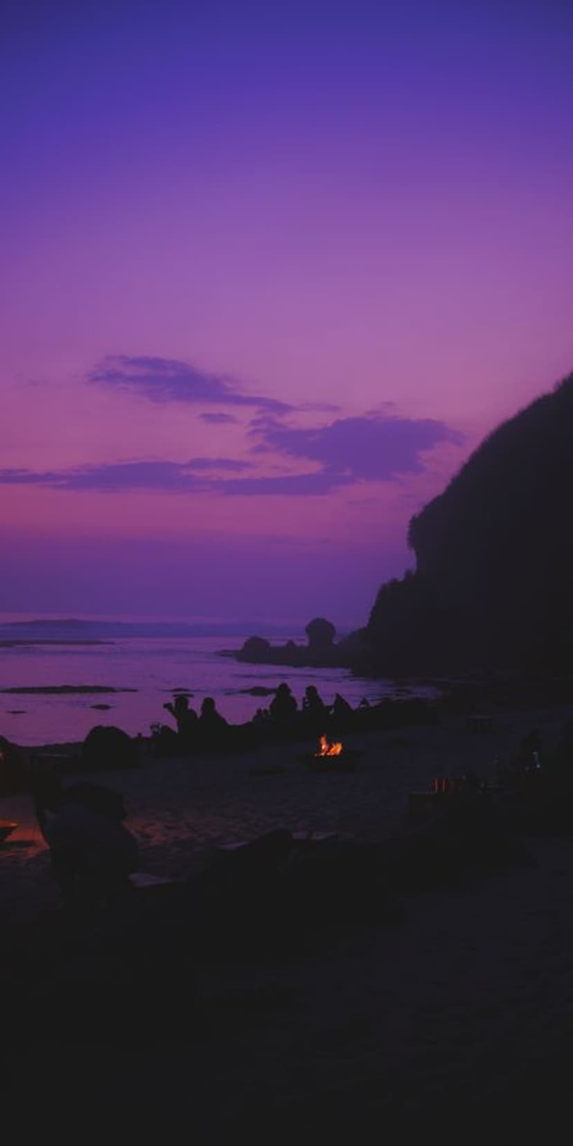 Glowing beach bonfire on a rocky shore beneath a vivid purple twilight sky with tiny silhouettes of beachgoers.