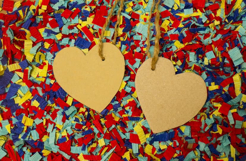Two white heart-shaped tags on twine hang above a dense layer of red, blue, and yellow circular paper confetti.
