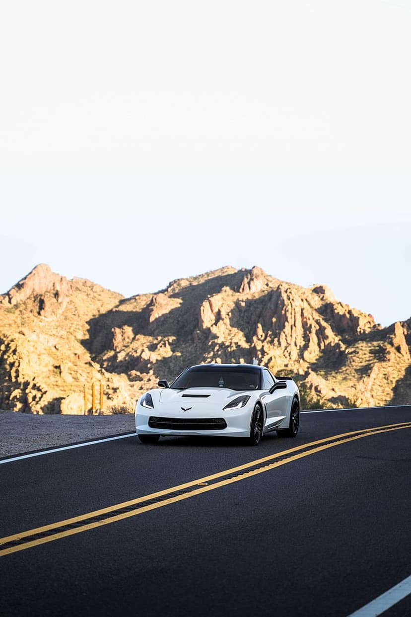 White Chevrolet Corvette driving on a winding asphalt road between rugged sun-drenched mountain peaks under blue sky.