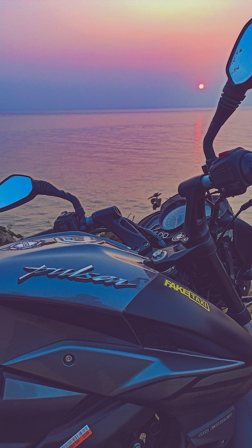 Pulsar motorcycle handlebars and fuel tank parked by a purple ocean sunset with pink sky reflections.