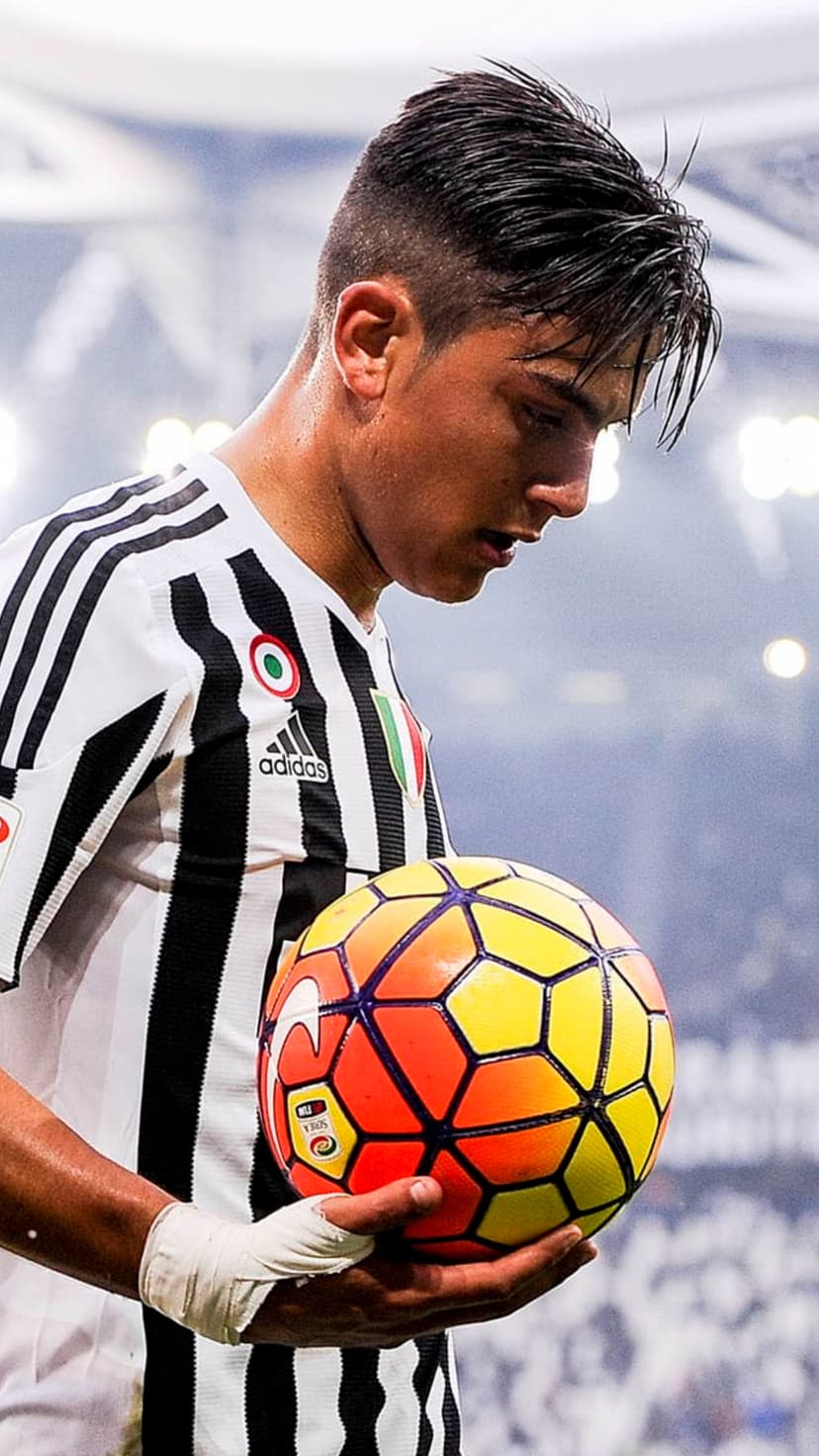 Paulo Dybala holds a bright orange soccer ball while wearing a black and white striped Juventus home jersey.