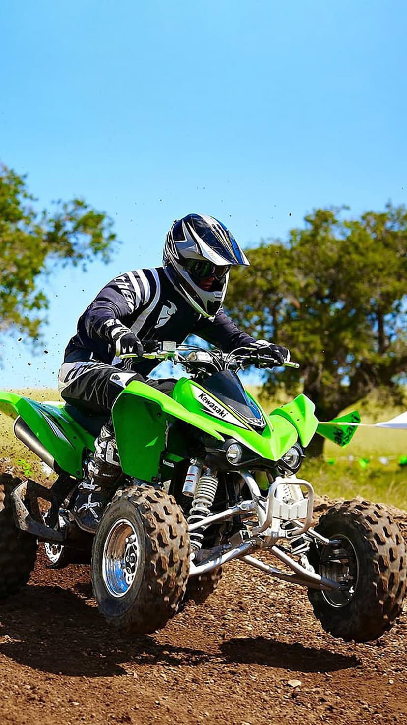 Kawasaki ATV rider in green racing gear kicks up a plume of brown dirt on a sunny outdoor track under a blue sky.