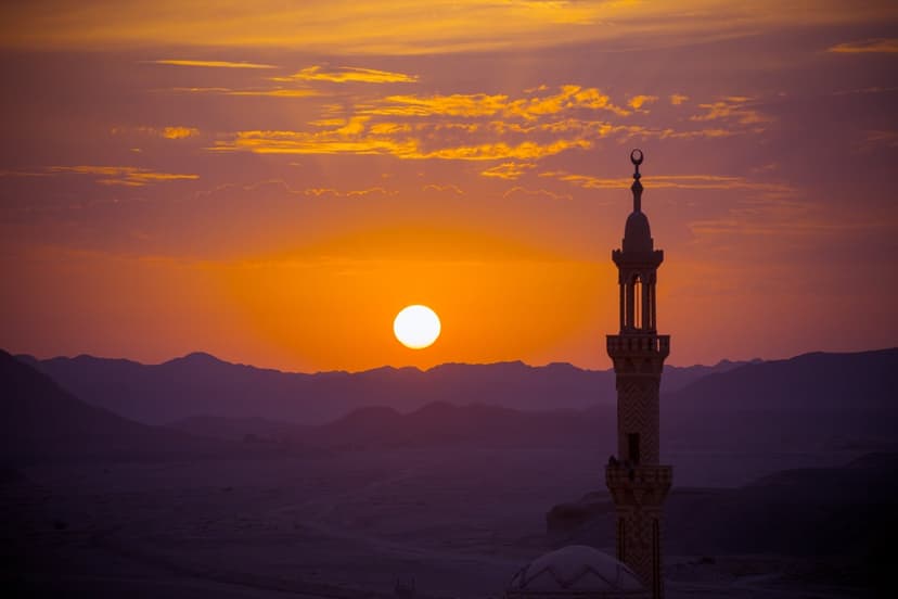 Mosque minaret stands beside purple desert mountains under a bright orange sun and golden clouds at sunset.