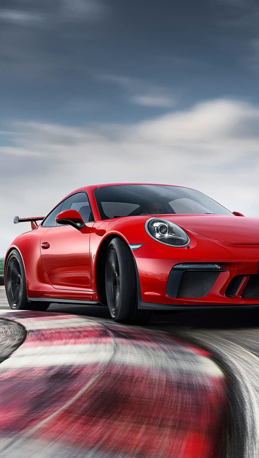 Porsche 911 GT3 R in red races through a track curve with blurred wheels against a bright cloudy sky.