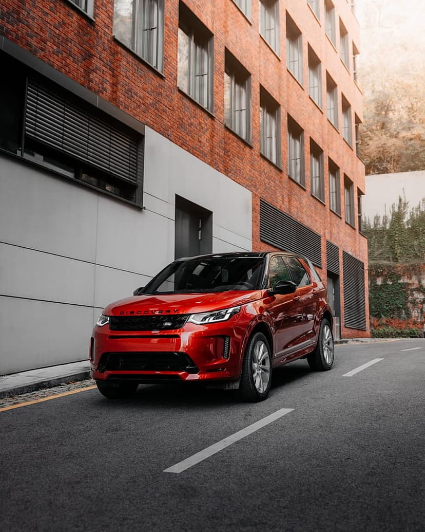 Red Land Rover SUV on a wet asphalt street before a contemporary brick facade with dark industrial louvers.