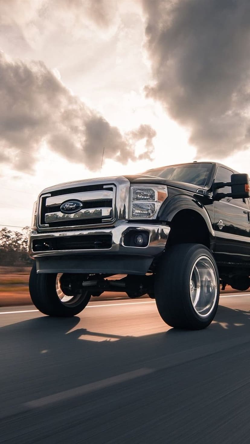 Black Ford F-Series Super Duty truck driving on an asphalt road with a chrome grille and dramatic cloudy sky.