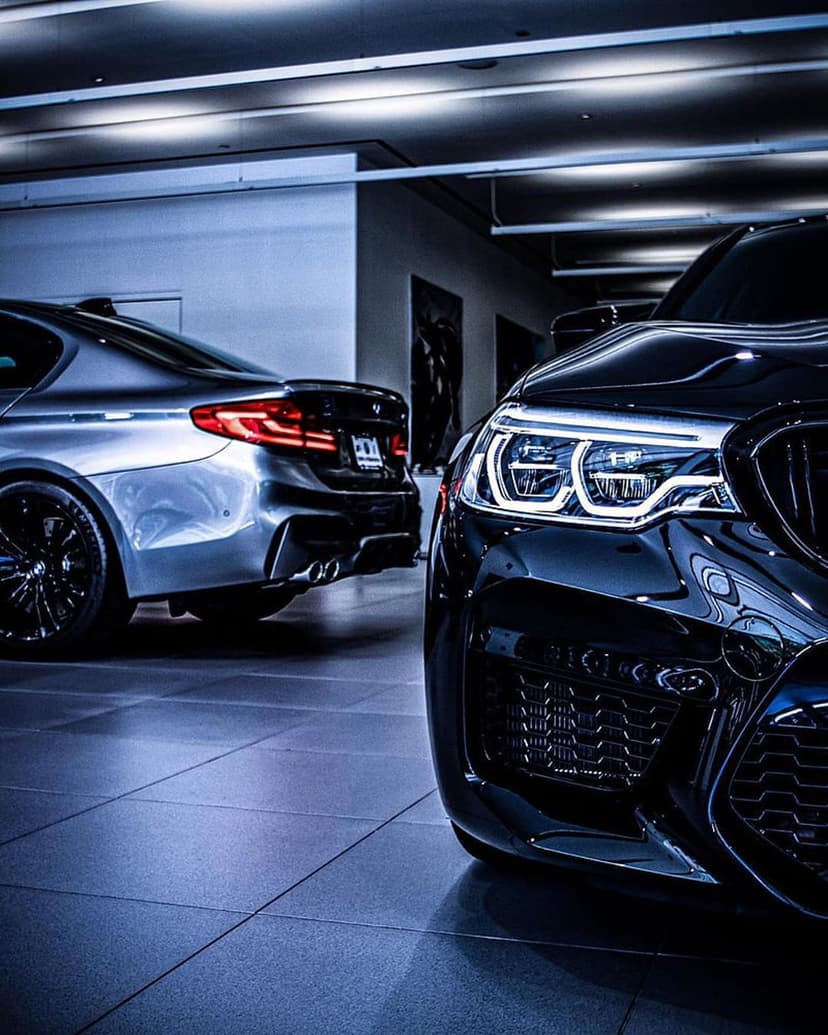 Silver sedan with a chrome grille parked next to a black luxury car under dramatic showroom ceiling lights.