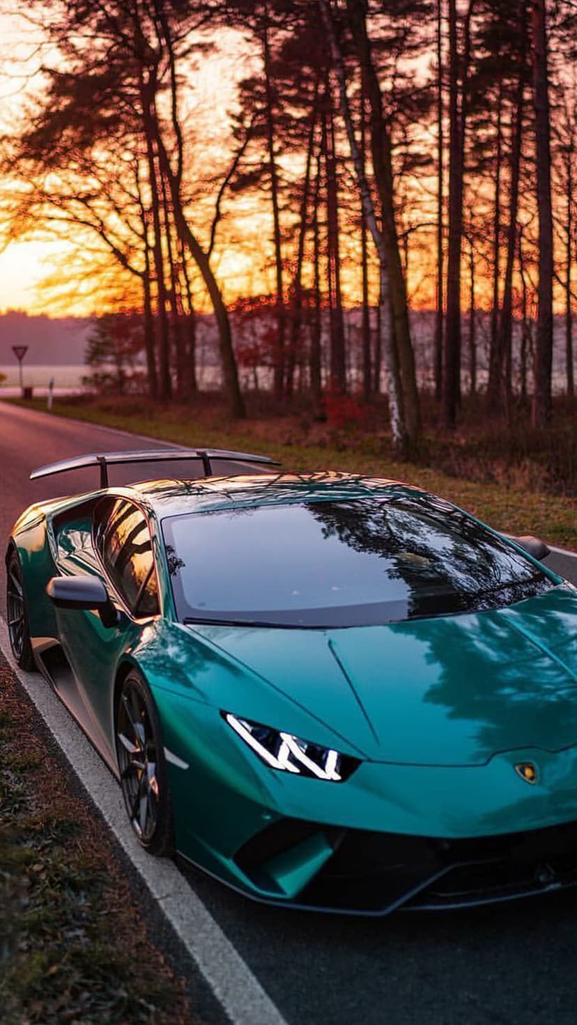Emerald green Lamborghini driving on a tree-lined asphalt road under a glowing orange and yellow sunset sky.