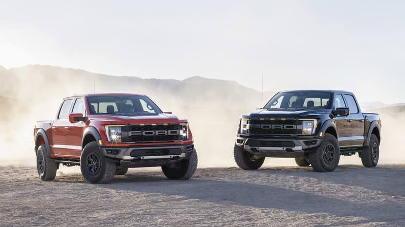 Orange and black Ford F-150 Raptor trucks parked in a dusty desert with mountains under a bright hazy sun.