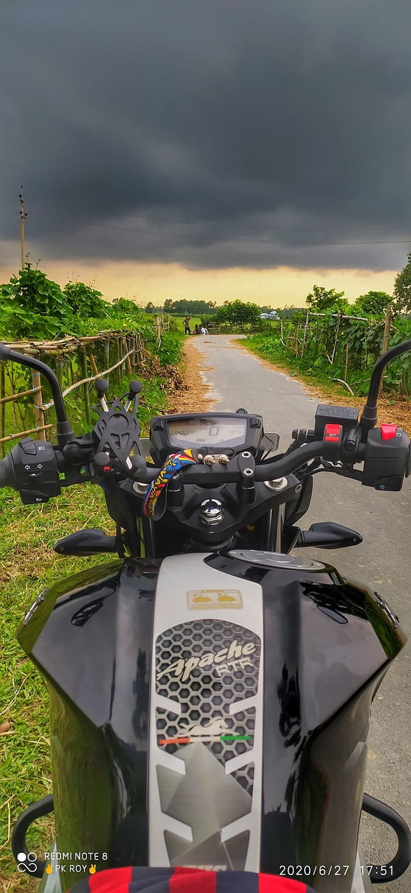 Apache RTR motorcycle handlebars and tank overlook a rural road beneath heavy dark storm clouds and green fields.