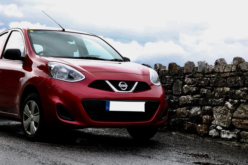 Red Nissan Micra parked on dark wet asphalt beside a rustic stone wall beneath a heavy, overcast moody sky.