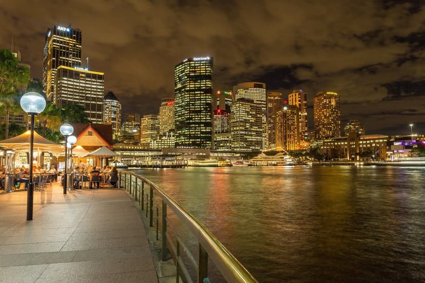 Sydney skyscrapers glow against a night sky with golden reflections shimmering on the water and a foreground pier.