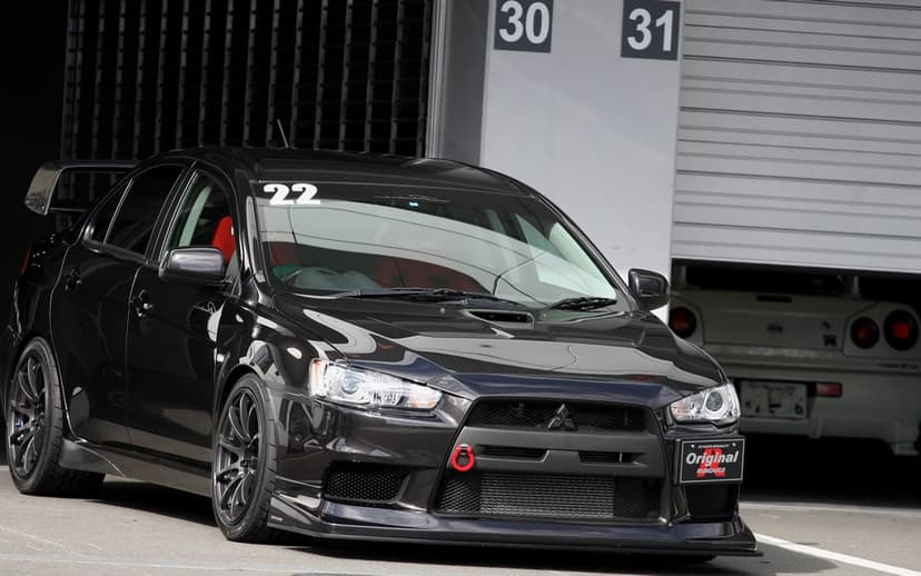 Black Mitsubishi Lancer Evolution X with red tow hook parked in a garage pit lane with a white R34 GT-R.