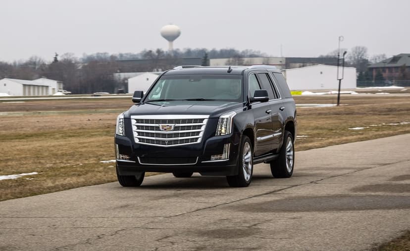 Dark blue Cadillac Escalade driving on an asphalt track under an overcast sky with chrome details visible.