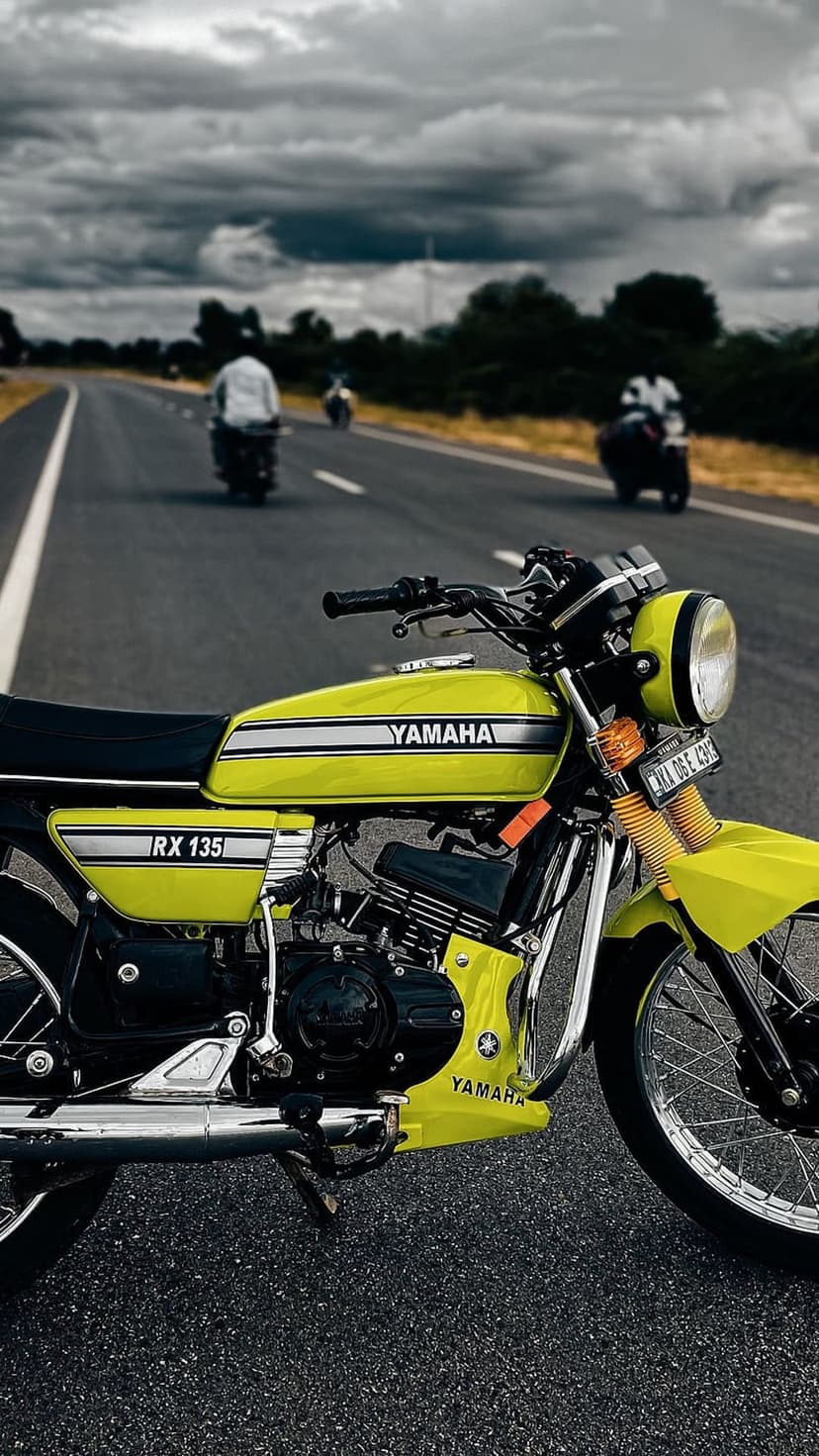 Yellow Yamaha RX 135 motorcycle on a dark asphalt road with a dramatic cloudy sky and distant riders.
