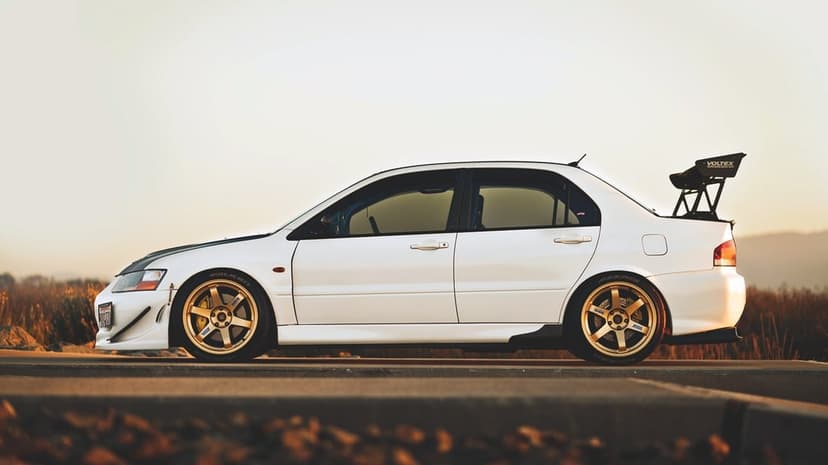 White Mitsubishi Lancer Evolution with gold wheels and a large wing parked on a road at sunset.